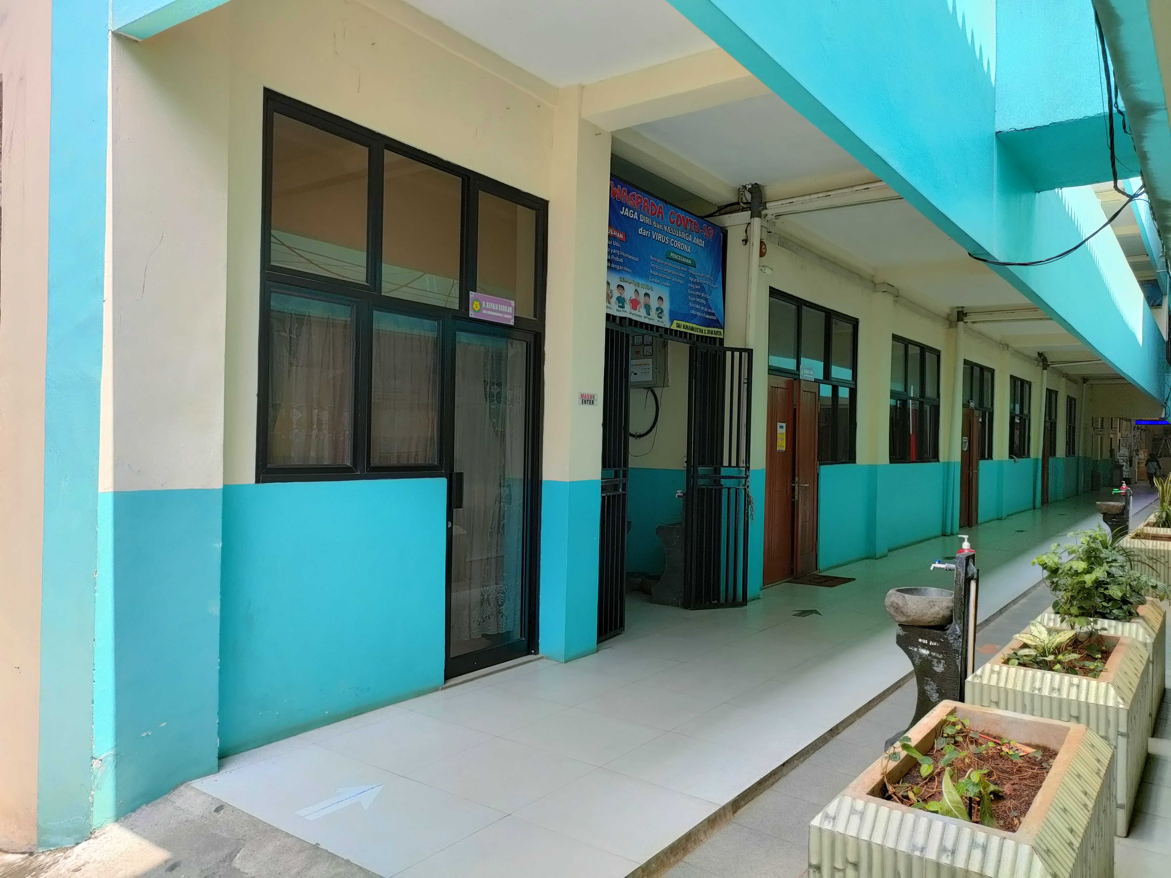 Koridor kelas berdinding krem dan biru toska dengan deretan pintu dan jendela berkusen hitam; papan nama Ruang Kepala Sekolah, poster imbauan kesehatan bertuliskan SMA Muhammadiyah 2 Jakarta, wastafel cuci tangan, dan bak tanaman berjajar di tepi lorong