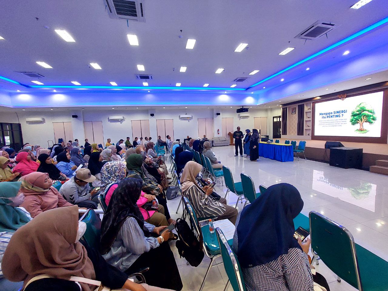 Aula sekolah berlantai keramik mengilap dengan lampu tersembunyi biru di plafon, panggung dan layar proyeksi di ujung ruangan, serta deretan kursi terisi peserta pertemuan