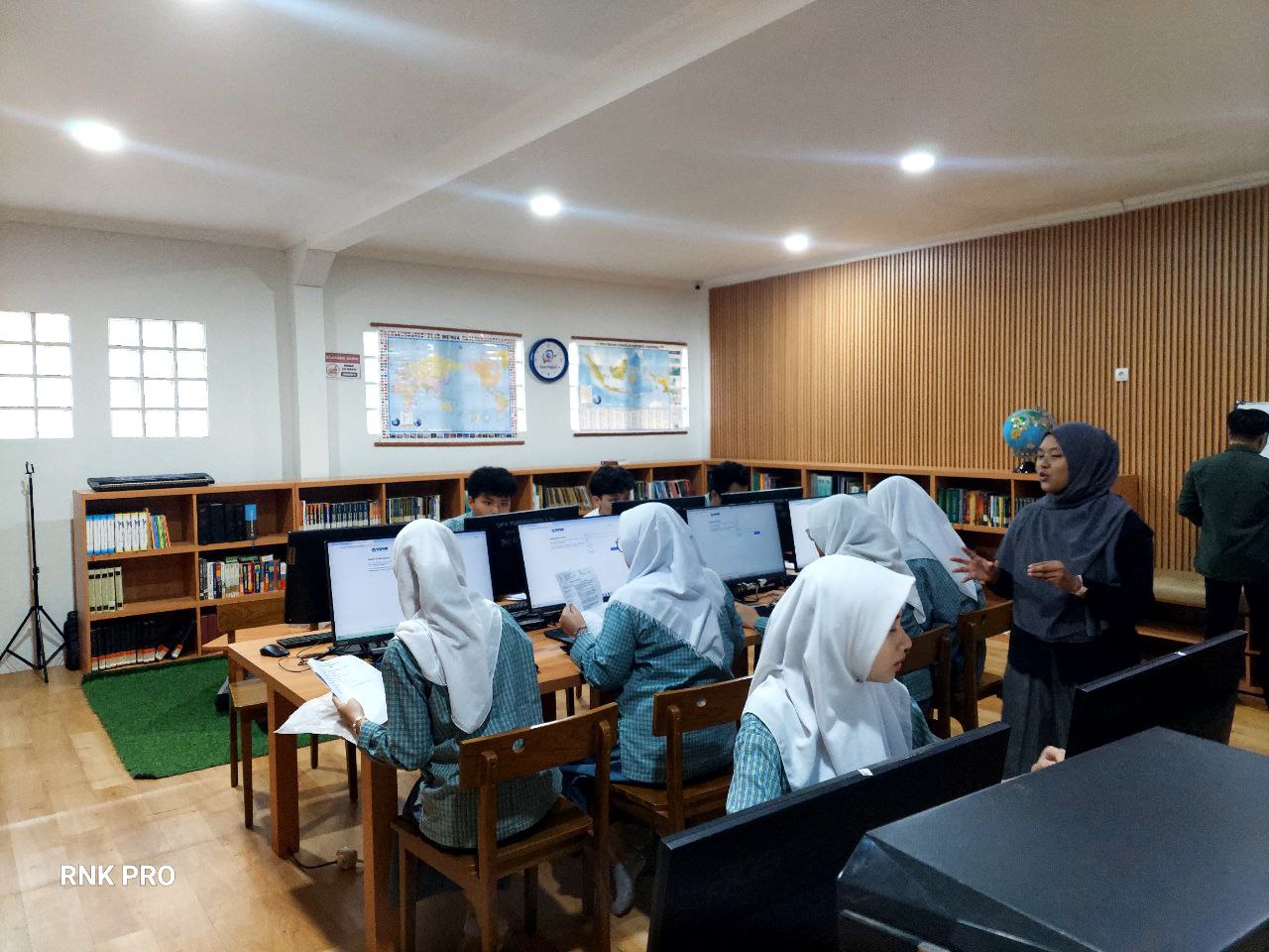Ruang perpustakaan sekaligus ruang komputer: siswa berseragam kotak-kotak biru duduk di meja kayu menghadap monitor, guru berdiri mendampingi, dengan rak buku sepanjang dinding, peta dunia, peta Indonesia, dan bola dunia
