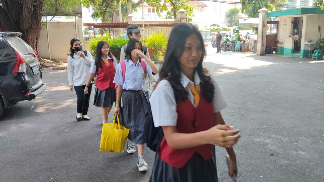 Pelataran sekolah berpaving dengan siswa berseragam putih, rompi maroon, dan rok abu berjalan keluar; pagar besi, pohon rindang, dan bangunan berkanopi hijau tampak di latar