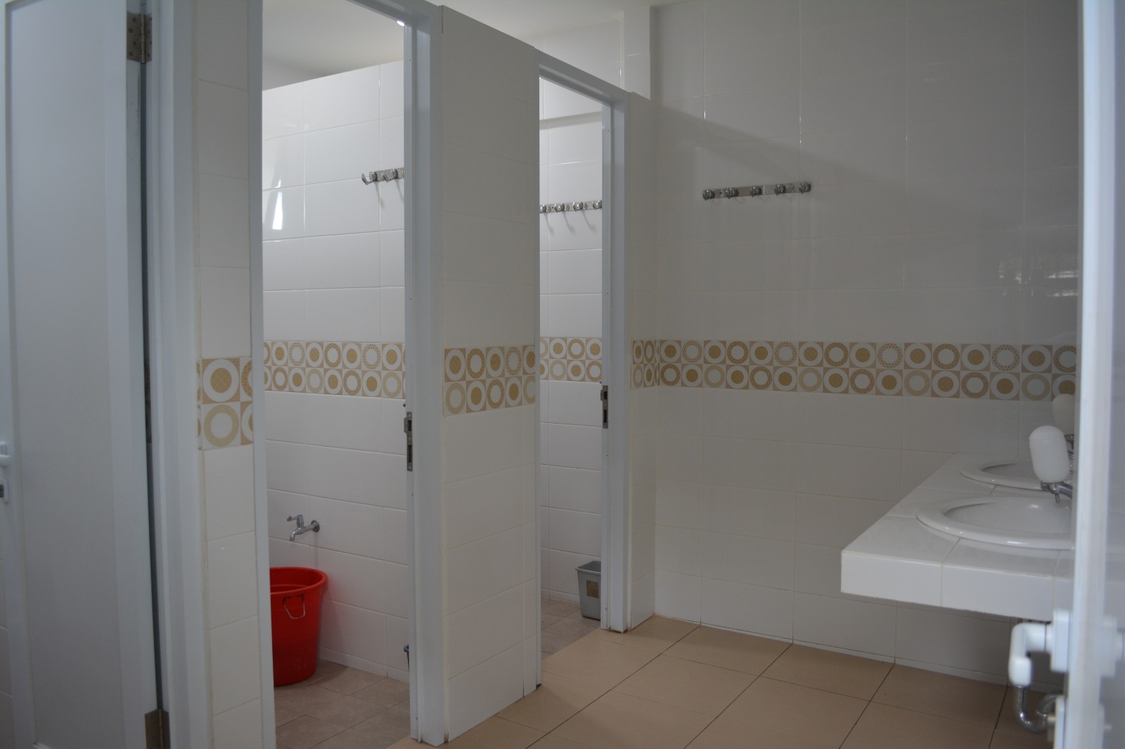 Kamar mandi sekolah berdinding keramik putih bermotif, dua bilik berpintu, gantungan baju, ember, dan meja wastafel ganda di sisi kanan
