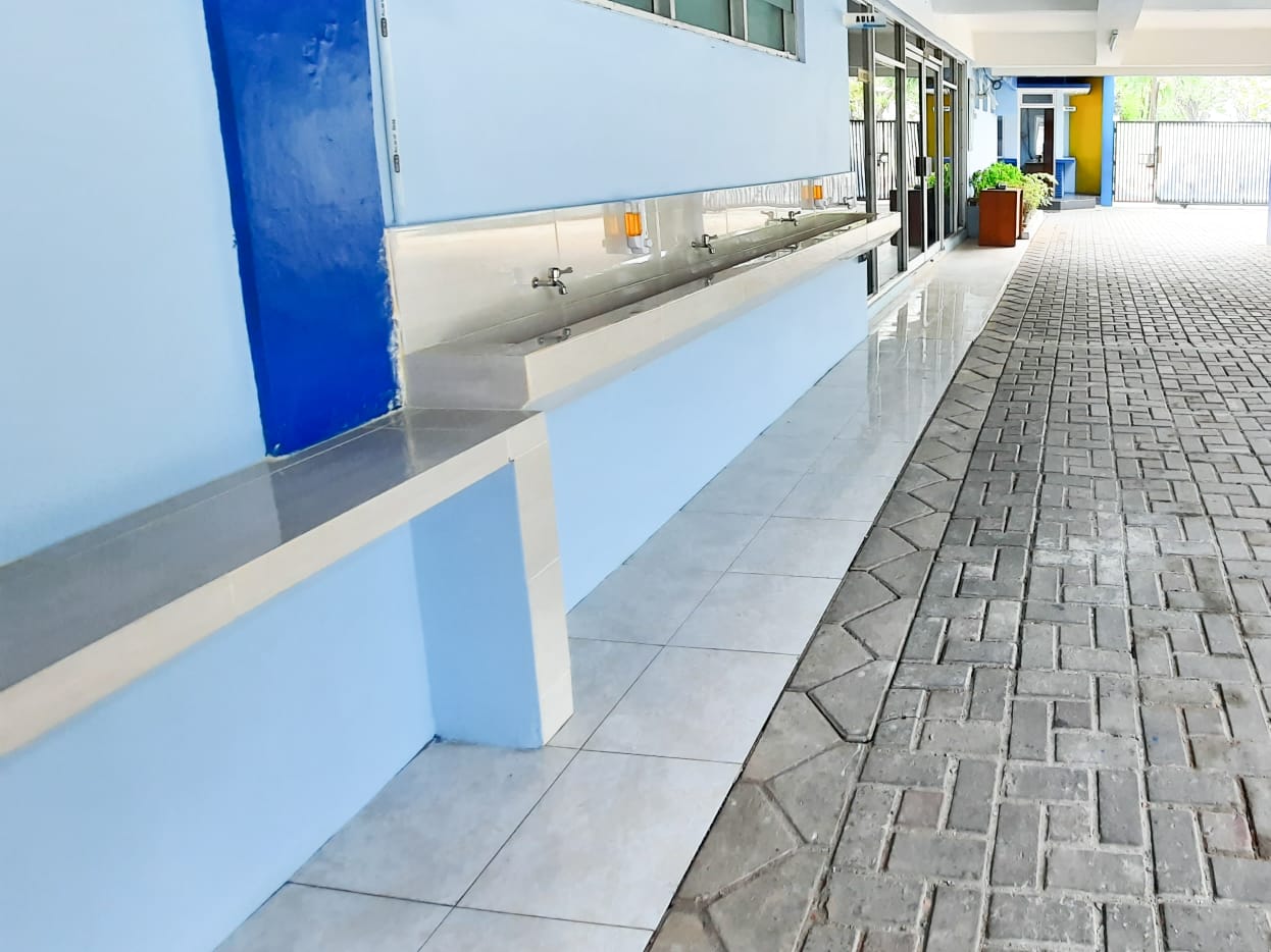 Deretan wastafel stainless memanjang di selasar beratap dengan beberapa keran dan botol sabun, menghadap jalan berpaving menuju pintu aula
