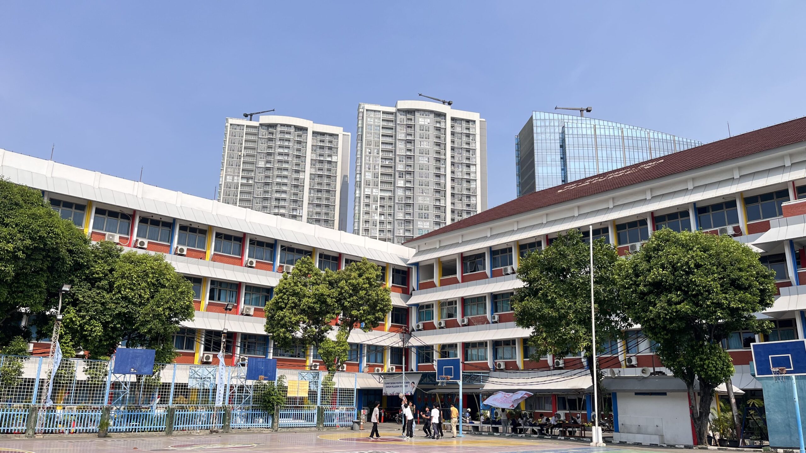 Dua sayap gedung kelas tiga lantai berkusen jendela merah, kuning, dan biru mengelilingi halaman berlapangan basket; siswa bermain di lapangan, pohon rindang di tepinya, dan gedung bertingkat tinggi tampak di kejauhan
