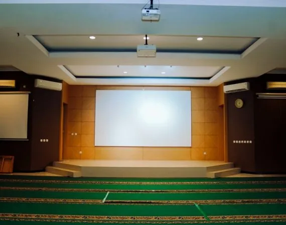 Aula berkarpet sajadah hijau menghadap panggung berlayar proyeksi, dua proyektor tergantung di langit-langit