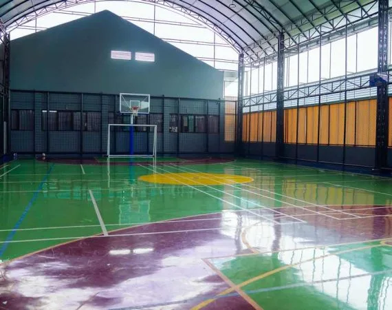 Lapangan beratap lengkung tanpa orang; garis basket dan futsal bertumpuk di lantai hijau-merah, dinding berjaring kawat di kedua sisi