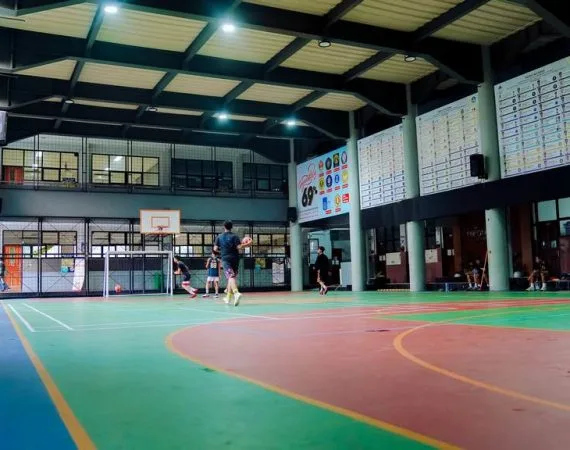 Lapangan indoor beratap baja dengan ring basket dan gawang futsal; sejumlah siswa sedang bermain, papan pengumuman dan spanduk angka 69 terpasang di dinding