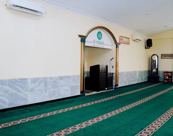 Ruang dalam masjid berkarpet hijau; papan nama bertuliskan Al-Hidayah dan Jakarta Timur terpasang di atas pintu lengkung