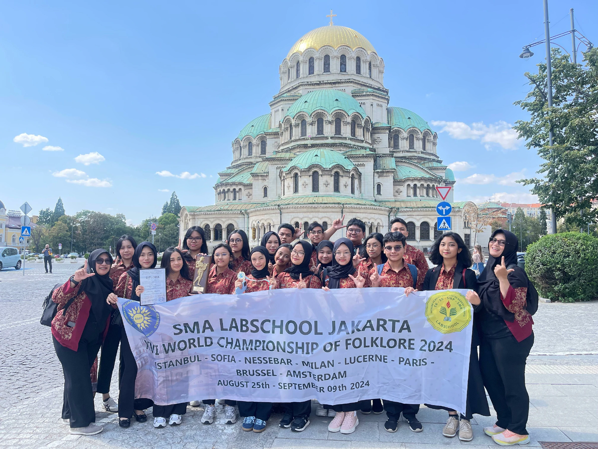Regu folklore sekolah berbusana batik merah membawa piala dan piagam di depan spanduk 'SMA Labschool Jakarta - XVI World Championship of Folklore 2024', dengan Katedral Alexander Nevsky di latar
