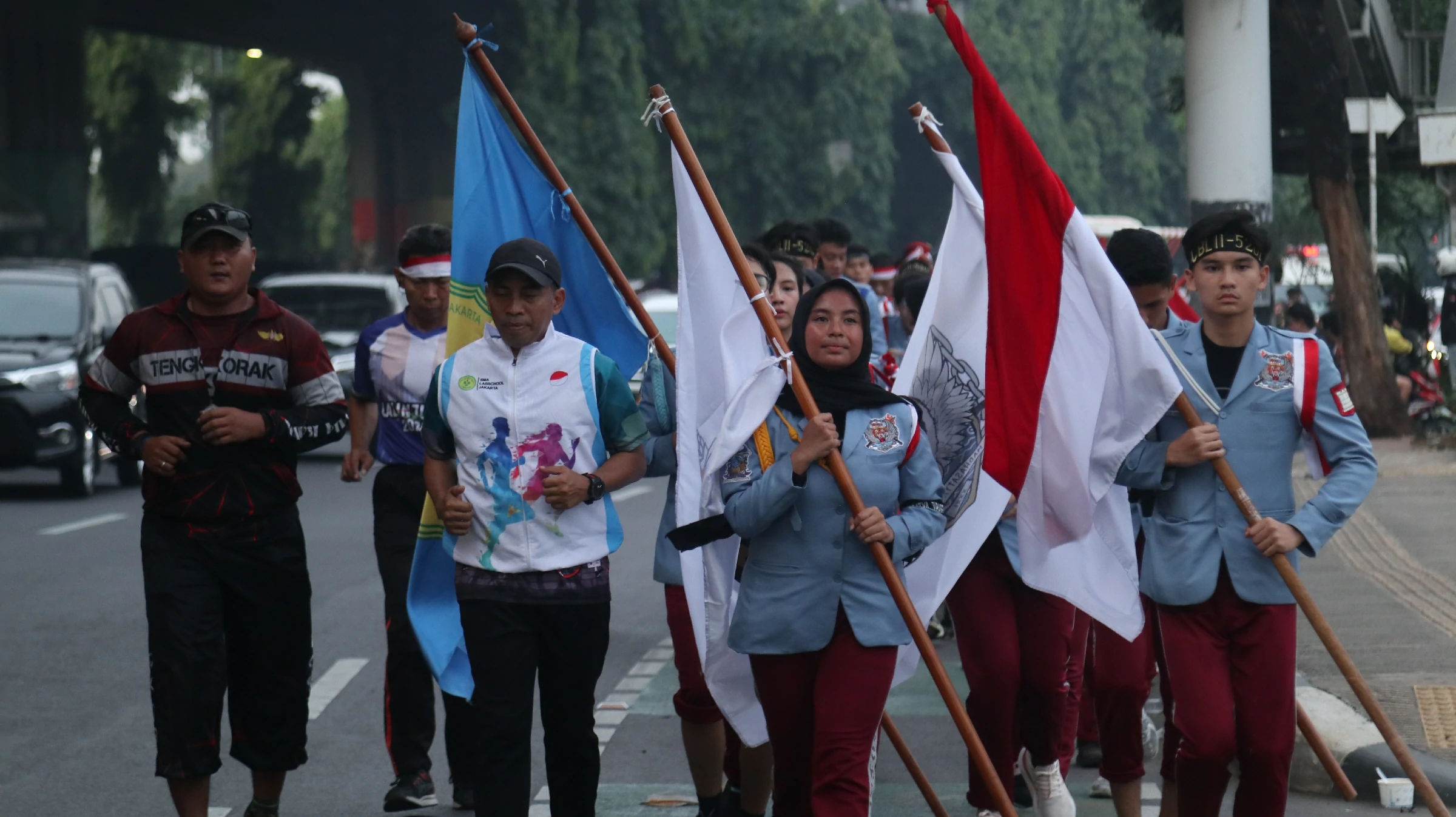 Barisan siswa berjas biru muda berlambang sekolah membawa bendera merah putih dan panji dalam kirab di jalan raya, didampingi pembina