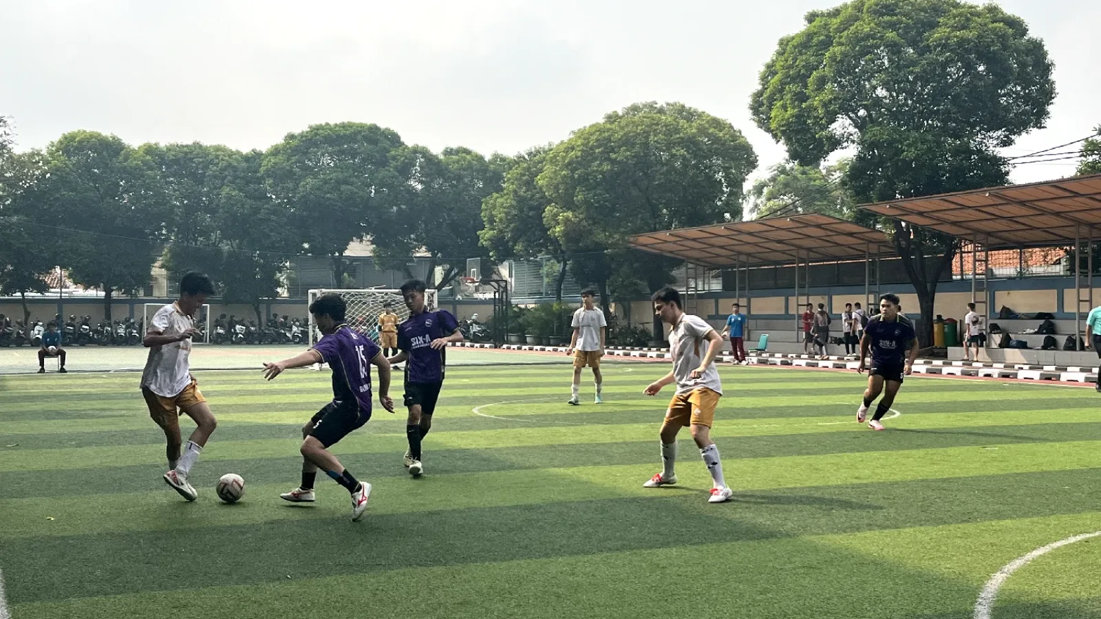 Lapangan rumput sintetis sekolah dengan siswa bermain sepak bola; tribun beratap, ring basket, dan pepohonan di sekeliling