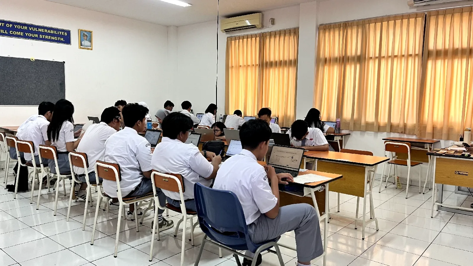 Ruang kelas berpendingin dengan siswa berseragam putih-abu mengerjakan tugas di laptop; papan tulis, meja kayu, dan spanduk motivasi berbahasa Inggris di dinding