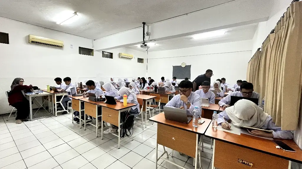 Ruang kelas berisi siswa berseragam putih di meja belajar kayu, sebagian memakai laptop dan tablet; proyektor, papan tulis, jam dinding, dan dua pendingin ruangan terlihat