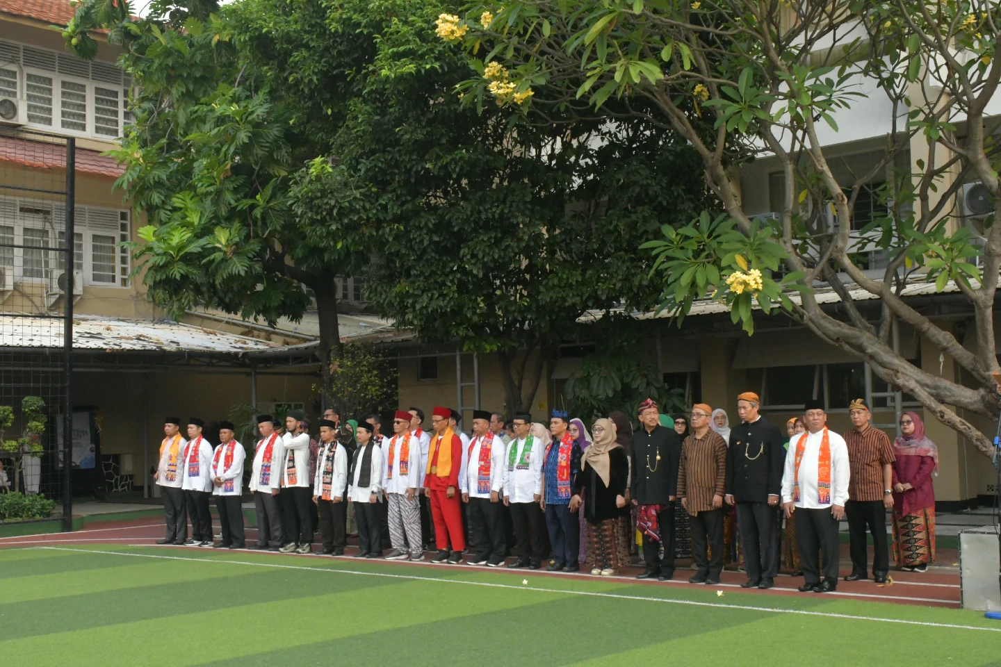 Guru dan tenaga kependidikan berbusana daerah berbaris di tepi lapangan sekolah, di bawah pohon; bangunan kelas bertingkat terlihat di kedua sisi