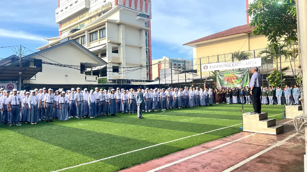 Upacara di lapangan rumput sintetis sekolah; barisan siswa berseragam putih-abu dan gedung sekolah terlihat di latar