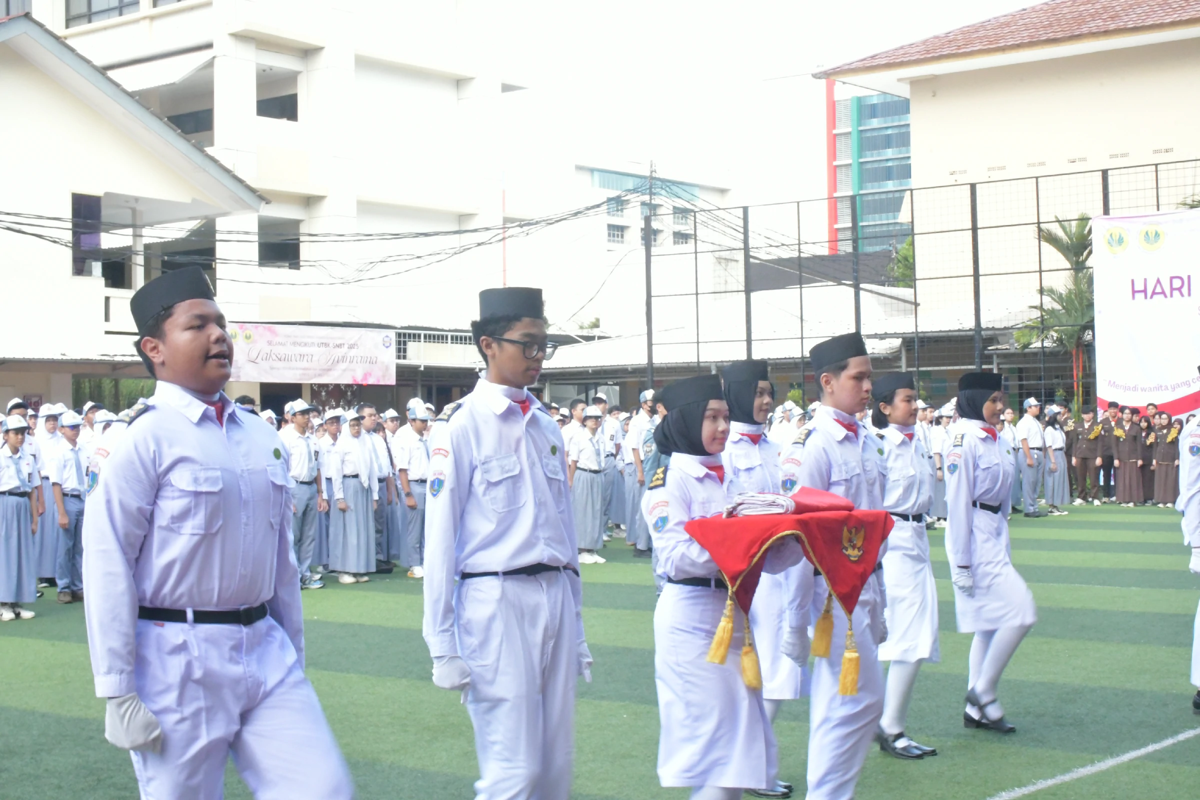 Regu pengibar bendera membawa bendera merah putih melintasi lapangan sekolah saat upacara, disaksikan barisan siswa; gedung kelas dan spanduk sekolah tampak di belakang