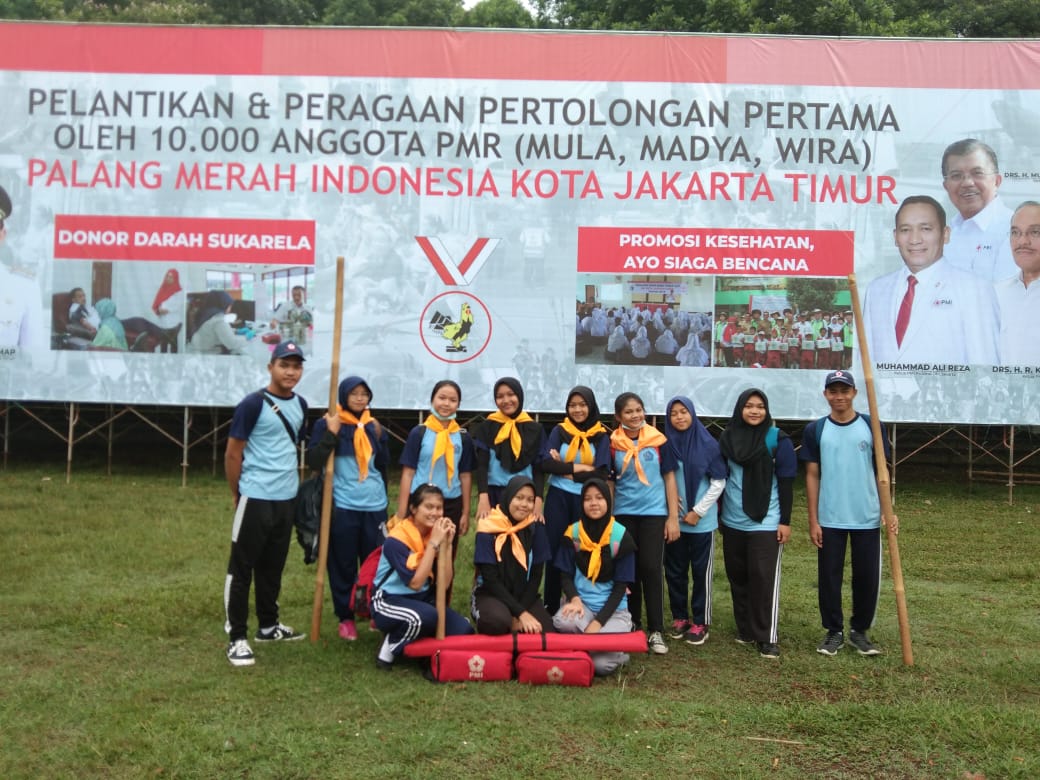 Regu Palang Merah Remaja berseragam olahraga sekolah dengan slayer oranye berfoto di lapangan rumput, di depan spanduk pelantikan dan peragaan pertolongan pertama Palang Merah Indonesia Kota Jakarta Timur