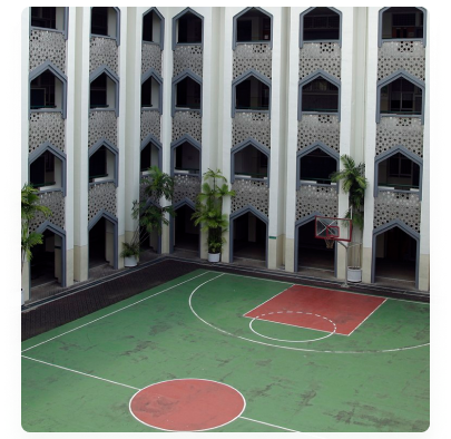 Lapangan basket beralas hijau merah di antara dua gedung kelas bertingkat berfasad krawangan putih, dengan tanaman pot di sisi lapangan