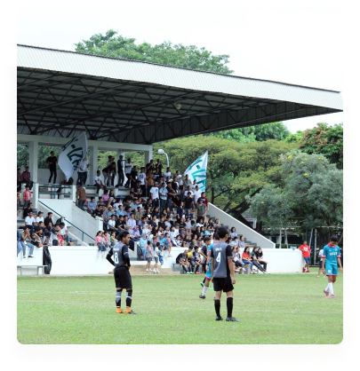 Lapangan sepak bola berumput dengan tribun beratap penuh penonton yang mengibarkan bendera, dan pemain berkaus hitam serta biru di tengah lapangan