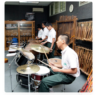 Ruang musik: siswa memainkan dua set drum, dengan deretan angklung tergantung di rak sepanjang dinding dan lemari alat musik di belakangnya