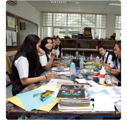 Ruang seni rupa: siswa mengerjakan karya di meja panjang penuh kertas warna, cat, kuas, dan gunting, dengan papan pajang karya di dinding