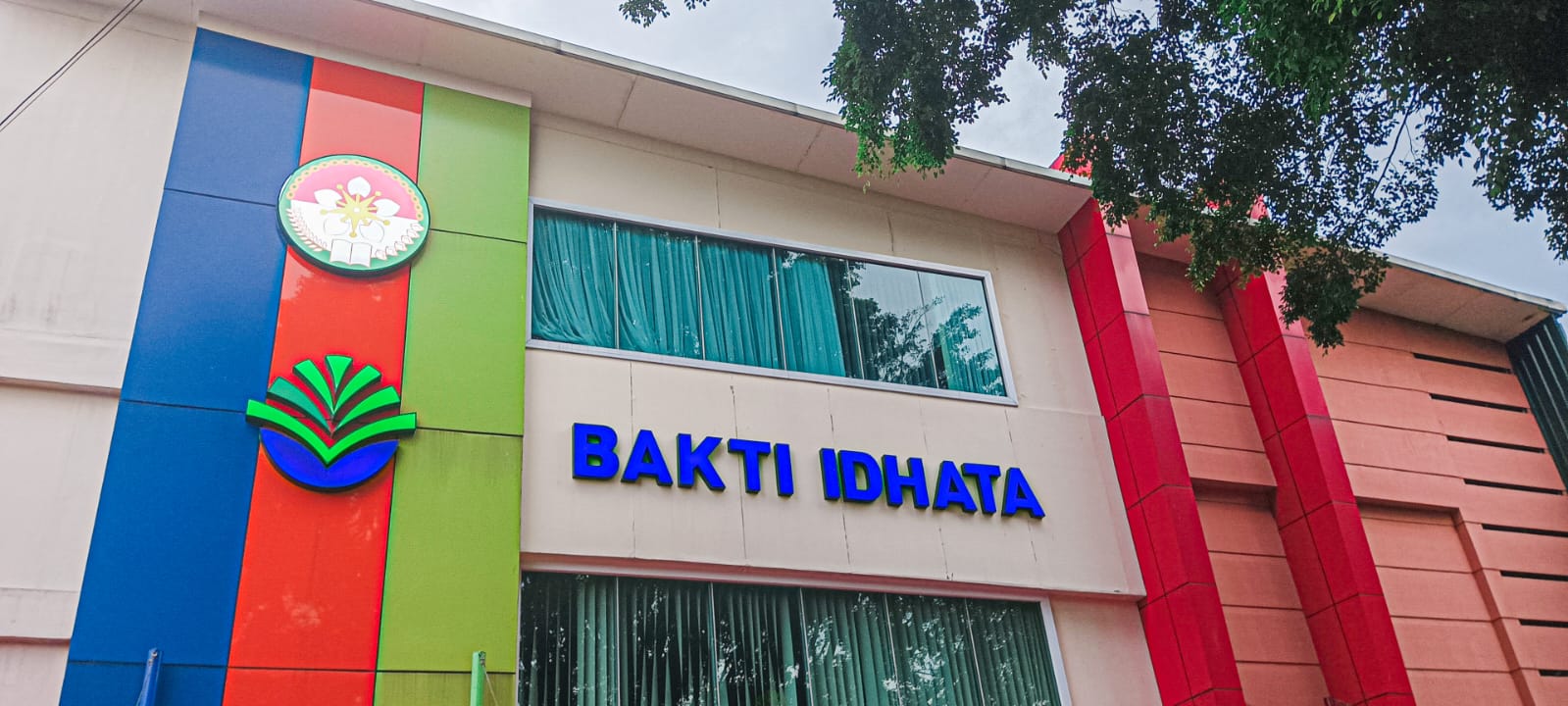 Fasad depan gedung sekolah bertuliskan 'BAKTI IDHATA' dengan dua lambang timbul, dinding berbidang biru, merah, dan hijau, serta jendela berkusen lebar