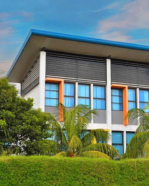 Gedung sekolah bertingkat dua dengan kisi-kisi kayu dan kolom jingga, dilihat dari sisi taman berumput.