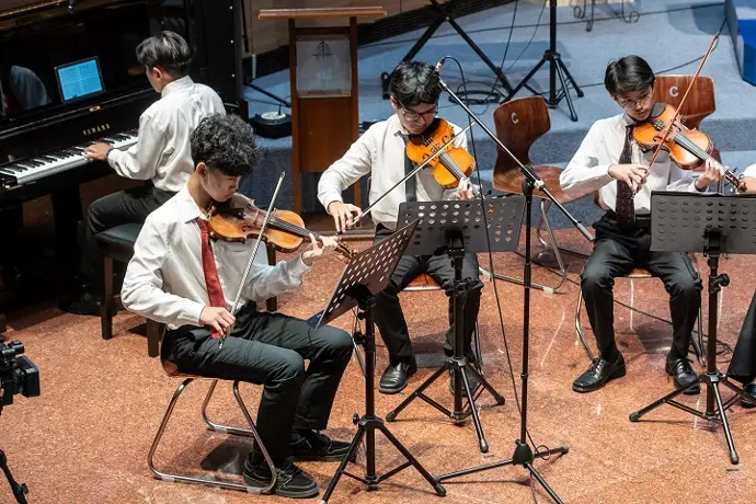 Siswa berkemeja putih dan berdasi memainkan biola bersama pengiring piano di atas panggung, lengkap dengan penyangga partitur dan mikrofon
