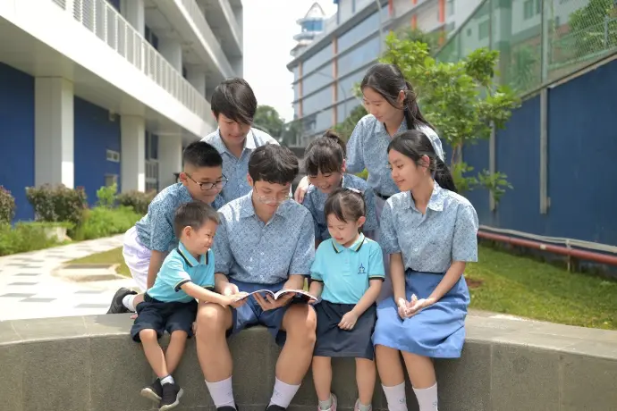 Sekelompok siswa berbagai jenjang duduk membaca di tepi taman halaman sekolah; gedung kelas bertingkat, koridor terbuka, dan pagar biru tampak di latar
