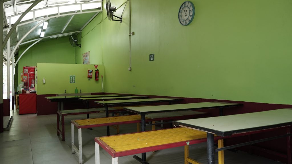 Kantin sekolah berdinding hijau dengan deretan meja panjang dan bangku kayu kuning, kipas dinding, jam dinding, dan lemari pendingin minuman di ujung