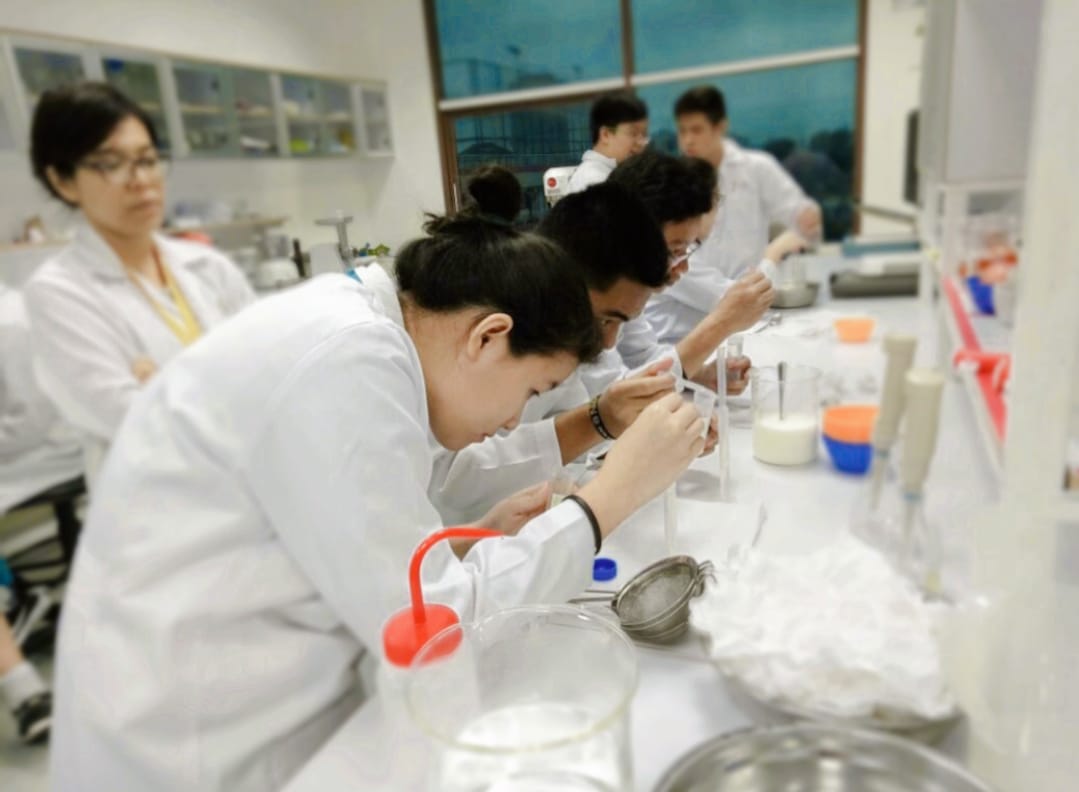 Laboratorium sains: siswa berjas lab putih memipet larutan ke dalam tabung di meja panjang berisi gelas ukur, saringan, dan botol reagen, dengan guru mendampingi di ujung meja