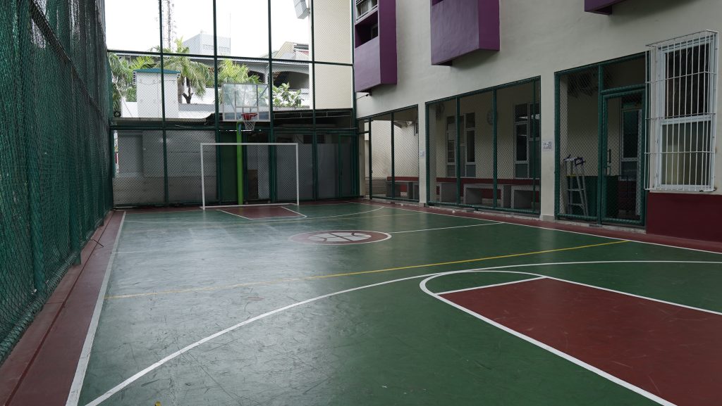 Lapangan serbaguna beralas hijau merah dengan ring basket dan gawang futsal, dipagari jaring tinggi, bersebelahan langsung dengan ruang kelas berjeruji