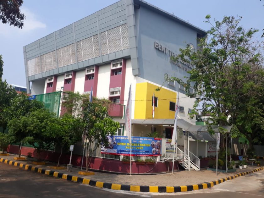 Gedung sekolah bertingkat berwarna biru, merah, dan kuning dilihat dari jalan masuk, bertuliskan 'Bent Tree School'; spanduk di pagarnya menyebut Perguruan Bina Maju Bangsa jenjang SMP dan SMA