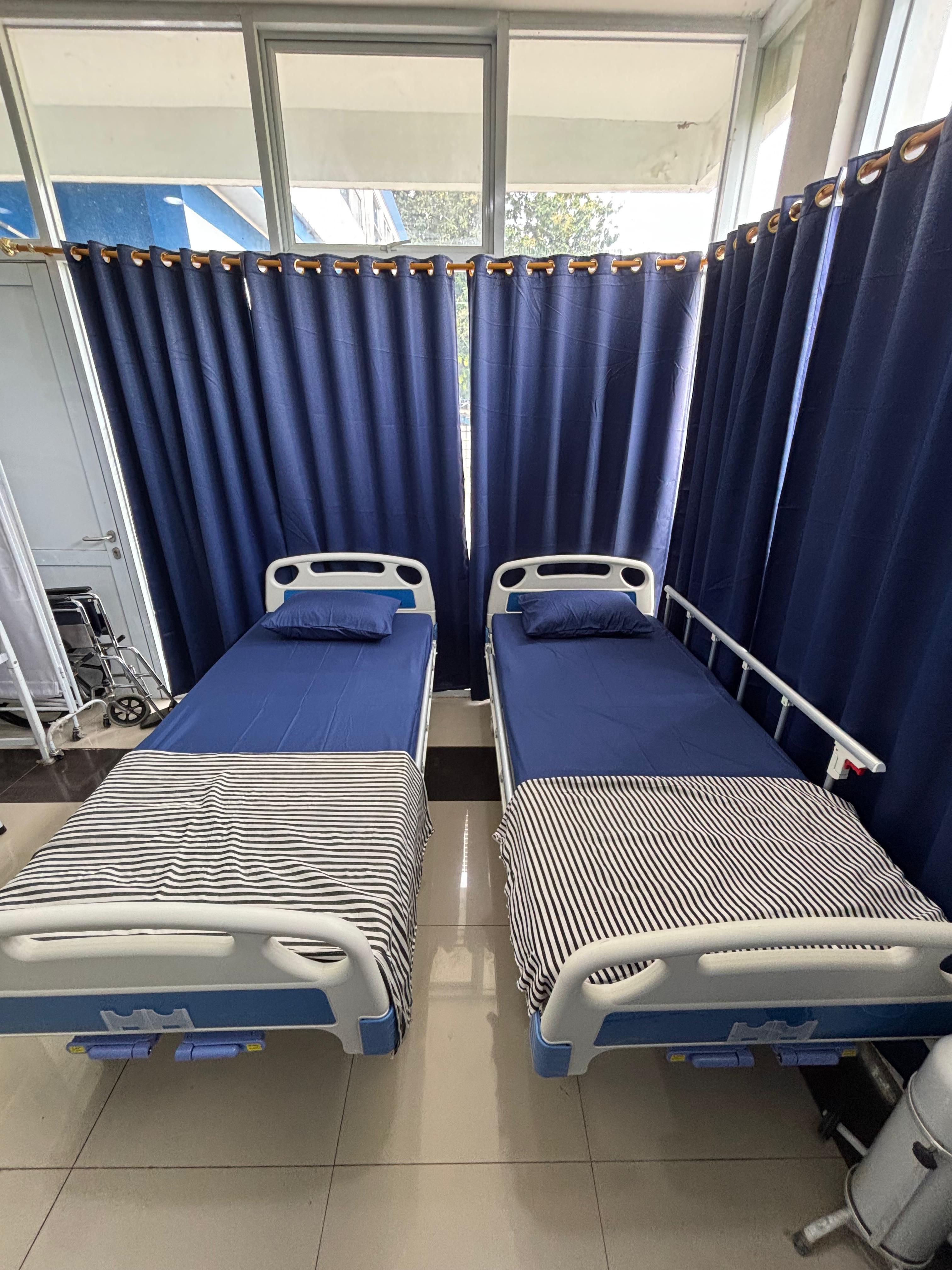 Ruang UKS berisi dua ranjang periksa beroda dengan sprei biru dan selimut bergaris, tirai privasi biru tua, serta kursi roda dan tiang infus di sudut ruangan